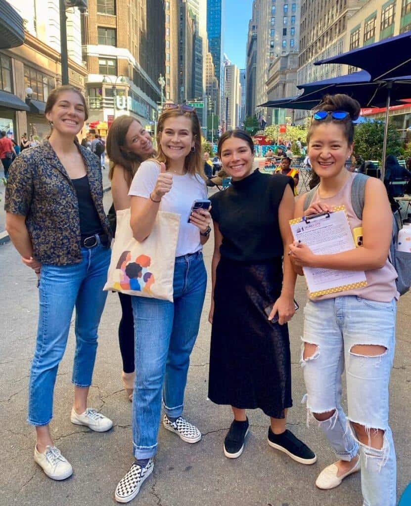 Team of five smiling participants on West 34th Street during a Herald Square historic scavenger hunt, holding a clipboard and phone with Midtown Manhattan skyscrapers in the background