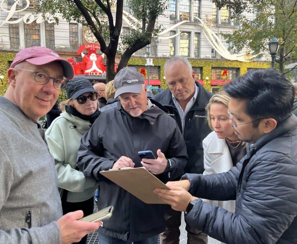 Corporate team reviewing clues on a clipboard and phone outside Macy’s Herald Square on 34th Street, surrounded by holiday decorations in Midtown Manhattan