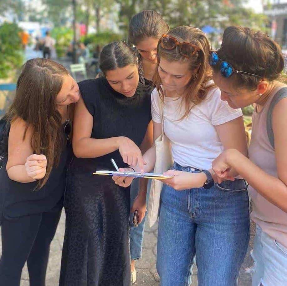 Team of participants huddled over a clipboard solving clues during a scavenger hunt in Bowling Green Park, Lower Manhattan